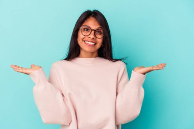 Mavi arka planda izole edilmiş genç Latin kadın kollarıyla ölçeği yapıyor, mutlu ve kendinden emin hissediyor..