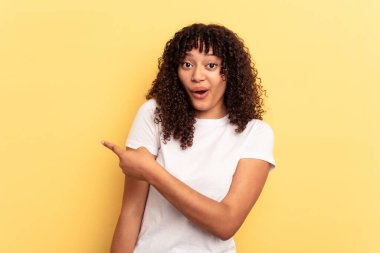 Sarı arka planda izole edilmiş genç melez kadın gülümsüyor ve boş alanda bir şeyler gösteriyor..