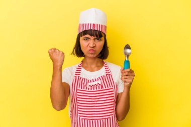 Genç melez dondurma üreticisi kadın elinde sarı arka planda izole edilmiş dondurma tutuyor. Genç melez dondurma üreticisi kadın sarı arka planda izole edilmiş bir dondurma kepçesiyle kameraya yumruğunu gösteriyor, agresif bir yüz ifadesi..