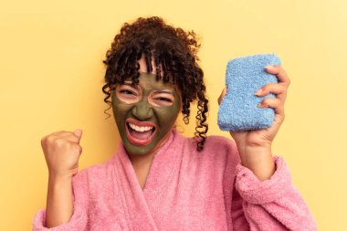 Pembe bornoz giyen, sarı arka planda izole edilmiş duş süngeri tutan genç bir ırk kadını zafer kazandıktan sonra yumruk kaldırıyor, kazanan konsept..