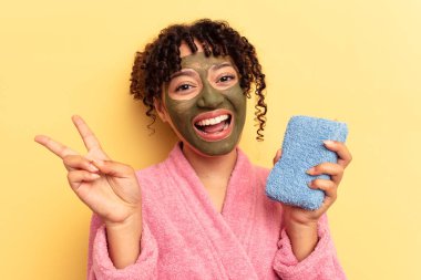 Pembe bornoz giyen genç bir ırk kadını sarı arka planda izole edilmiş bir duş süngeri tutuyor neşeli ve kaygısız parmaklarıyla barış sembolü gösteriyor..