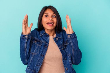 Mavi arka planda izole edilmiş genç Latin kadın hoş bir sürprizle karşılaşıyor, heyecanlı ve el kaldırıyor..