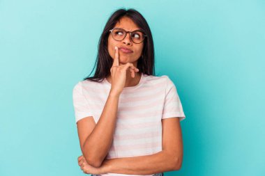 Mavi arka planda izole edilmiş genç Latin kadın şüpheli ve şüpheci bir ifadeyle yana bakıyor..