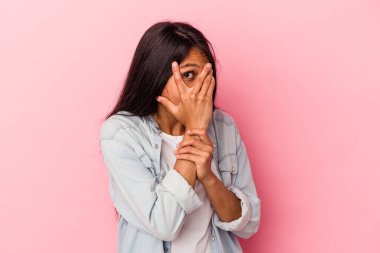 Pembe arka planda izole edilmiş genç Latin kadın parmaklarının arasından korkmuş ve gergin bir şekilde göz kırpıyor..