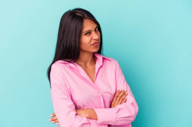 Mavi arka planda izole edilmiş, hoşnutsuzca kaşlarını çatan, kollarını kavuşturan genç Latin bir kadın..
