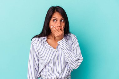 Genç Latin bir kadın mavi arka planda izole edilmiş. Düşünceli bir şekilde bir fotokopi odasına bakıyor. Ağzını eliyle kapatıyor..