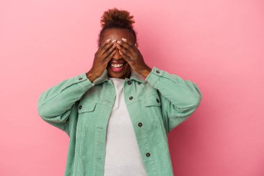Pembe arka planda izole edilmiş genç Afrikalı Amerikalı kadın. Gözleri ellerle kaplıyor. Gülümsüyor ve bir sürpriz bekliyor..