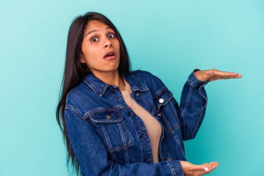 Mavi arka planda izole edilmiş genç Latin kadın iki eliyle bir şey tutuyor, ürün sunumu..