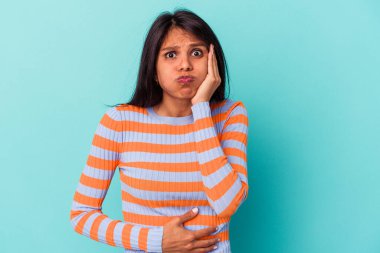 Mavi arka planda izole edilmiş genç Latin kadın yanakları patlatıyor, yorgun bir ifade takınıyor. Yüz ifadesi kavramı.