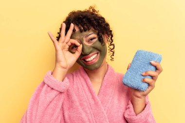 Pembe bornoz giyen genç bir ırk kadını sarı arka planda izole edilmiş duş süngeri tutuyor..