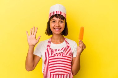 Küçük melez dondurma üreticisi kadın sarı arka planda izole edilmiş dondurma tutuyor. Beş numarayı parmaklarıyla gülümsüyor..