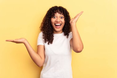 Sarı arka planda izole edilmiş genç melez kadın avuç içinde fotokopi alanı tutuyor, el üstünde tutuyor. Şaşırmış ve mutlu..