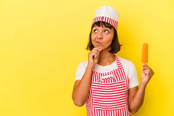 年轻的混血冰淇淋制造商女人拿着一个黄色背景的冰淇淋 侧着看 脸上带着怀疑和怀疑的表情 — 图库照片