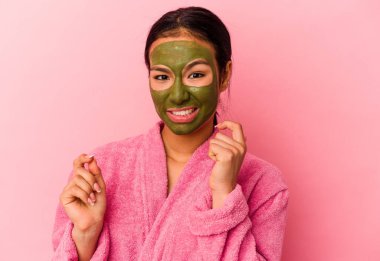 Genç Venezuelalı kadın, pembe arka planda izole edilmiş bornoz ve yüz maskesi takıyor tırnak yiyor, gergin ve endişeli..