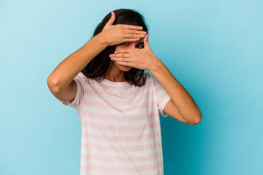 Mavi arka planda izole edilmiş genç beyaz kadın kameraya parmaklarıyla göz kırpıyor, yüzünü kapatmaktan utanıyor..