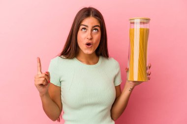 Elinde makarna kavanozu olan beyaz bir kadın pembe arka planda izole edilmiş ve ağzı açık bir şekilde üst tarafı işaret ediyor..