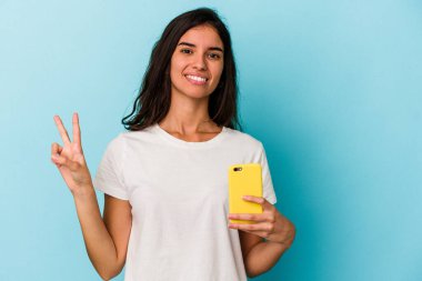 Elinde cep telefonu olan beyaz bir kadın mavi arka planda iki numarayı parmaklarıyla gösteriyor..