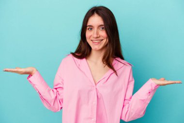 Mavi arka planda izole genç beyaz kadın kollarıyla ölçek yapıyor, mutlu ve kendinden emin hissediyor..