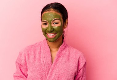 Genç Venezuelalı kadın bornoz ve yüz maskesi takıyor pembe arka planda izole edilmiş mutlu, güler yüzlü ve neşeli..