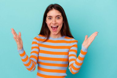 Mavi arka planda izole genç beyaz kadın hoş bir sürpriz alıyor, heyecanlı ve el kaldırıyor..