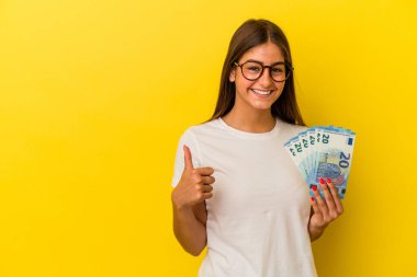 Sarı arka planda izole edilmiş banknotları tutan genç beyaz kadın gülümsüyor ve başparmağını kaldırıyor.