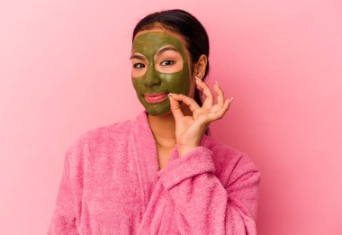 Bornoz ve yüz maskesi takan genç Venezuelalı kadın pembe arka planda izole edilmiş ve dudakları dudaklarında bir sır saklıyor..