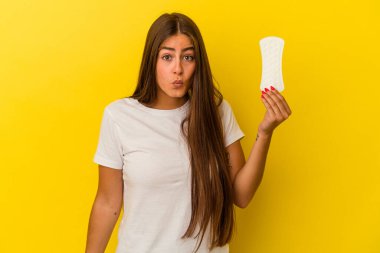 Sarı arka planda izole edilmiş bir kompres tutan genç beyaz kadın omuzlarını silker ve gözleri açık kalır..