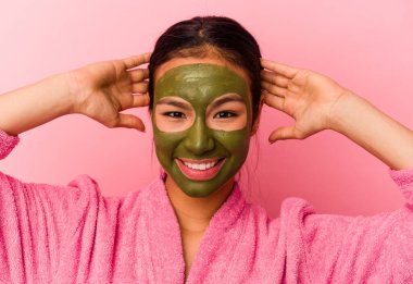 Genç Venezuelalı kadın, pembe arka planda yalıtılmış bornoz ve yüz maskesi giyiyor.