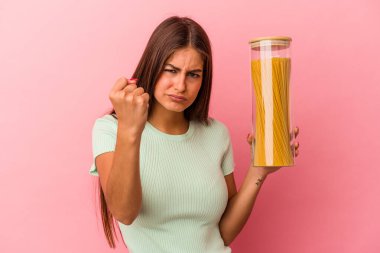 Elinde makarna kavanozu tutan beyaz bir kadın pembe arka planda izole edilmiş. Kameraya yumruğunu, agresif yüz ifadesini gösteriyor..