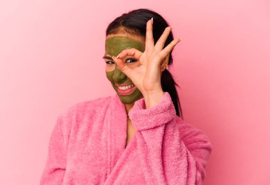 Genç Venezuelalı kadın, pembe arka planda izole edilmiş bornoz ve yüz maskesi takıyor..