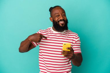 Elinde cep telefonu olan Afro-Amerikan bir adam mavi arka planda izole edilmiş bir şekilde gömlek fotokopi alanını gösteriyor, gururlu ve kendinden emin.