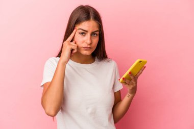 Elinde cep telefonu olan beyaz bir kadın pembe arka planda izole edilmiş bir şekilde tapınağı işaret ediyor, düşünüyor ve bir göreve odaklanıyor..