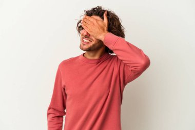 Beyaz arka planda soyutlanmış beyaz tenli genç bir adam elleriyle gözlerini kapatıyor, geniş bir gülümsemeyle bir sürpriz bekliyor..