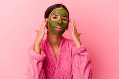 Pembe arka planda yalıtılmış bornoz ve yüz maskesi takan beyaz bir kadın.