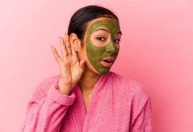 Genç Venezuelalı kadın, pembe arka planda izole edilmiş bornoz ve yüz maskesi takıyor ve bir dedikoduyu dinlemeye çalışıyor..