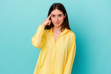 Mavi arka planda izole edilmiş genç beyaz kadın şakağı parmağıyla işaret ediyor, düşünüyor ve bir göreve odaklanıyor..