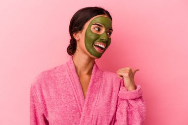 Bornoz ve yüz maskesi takan genç beyaz kadın pembe arka plan noktalarına izole edilmiş, baş parmak uzakta, gülen ve kaygısız..