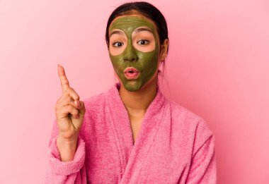 Genç Venezuelalı kadın, pembe arka planda izole edilmiş bornoz ve yüz maskesi takıyor. Harika bir fikri var, yaratıcılık kavramı..