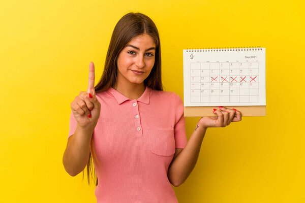 Young caucasian woman holding a calendar isolated on yellow background showing number one with finger.