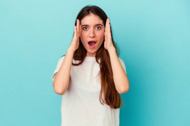 Mavi arka planda izole edilmiş genç beyaz kadın şaşırdı ve şok oldu..