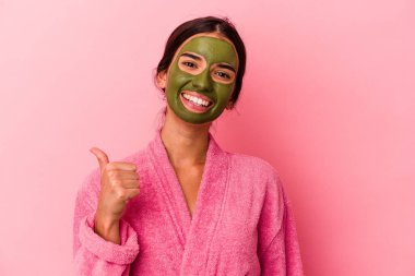 Pembe arka planda izole edilmiş bornoz ve yüz maskesi takan beyaz bir kadın gülümsüyor ve başparmağını kaldırıyor.