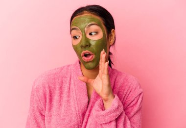 Genç Venezuelalı bir kadın pembe arka planda izole edilmiş bornoz ve yüz maskesi takıyor. Gizli bir sıcak fren haberi veriyor ve yan tarafa bakıyor.