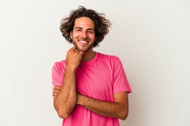 Beyaz arka planda izole olmuş genç beyaz bir adam gülümsüyor ve kendinden emin, çenesine elleriyle dokunuyor..