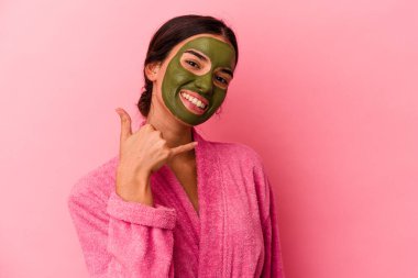 Pembe arka planda izole edilmiş bornoz ve yüz maskesi takan genç beyaz kadın parmaklarıyla bir cep telefonu hareketi gösteriyor..