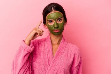 Pembe arka planda izole edilmiş bornoz ve yüz maskesi takan genç beyaz kadın şakağı parmağıyla işaret ediyor, düşünüyor ve bir göreve odaklanıyor..