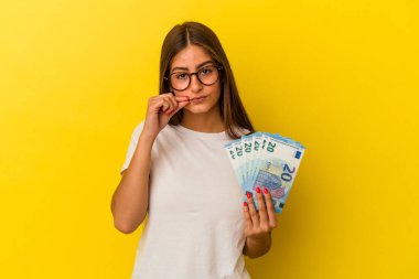 Sarı arka planda izole edilmiş banknotlar tutan genç beyaz kadın dudaklarında parmaklar ile bir sır saklıyor..