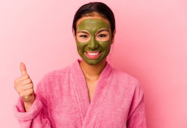 Genç Venezuelalı kadın pembe arka planda izole edilmiş bornoz ve yüz maskesi giyiyor gülümsüyor ve başparmağını kaldırıyor.