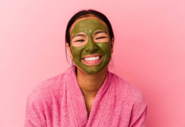 Genç Venezuelalı kadın, pembe arka planda izole edilmiş bornoz ve yüz maskesi giyip gülüyor ve eğleniyor..