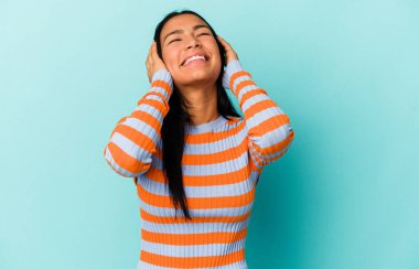 Genç Venezuelalı bir kadın mavi arka planda izole edilmiş, mutlu bir şekilde ellerini başının üstünde tutarak gülüyor. Mutluluk kavramı.