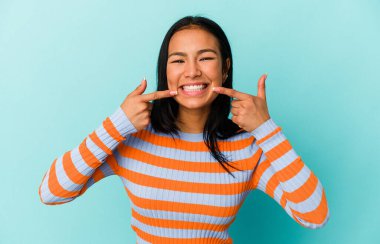 Genç Venezuelalı bir kadın mavi arka planda izole edilmiş gülümsüyor, parmaklarını ağzına doğrultuyor..